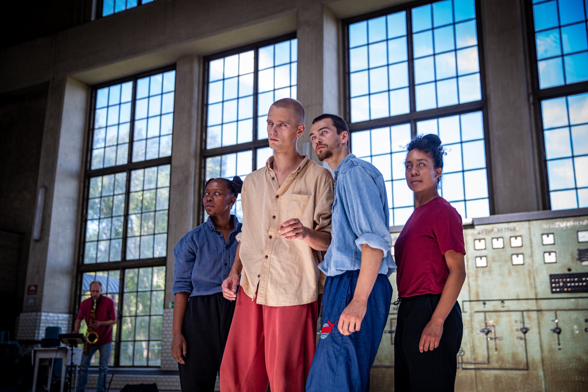 Four dancers standing closely together in a sunlit industrial rehearsal space.