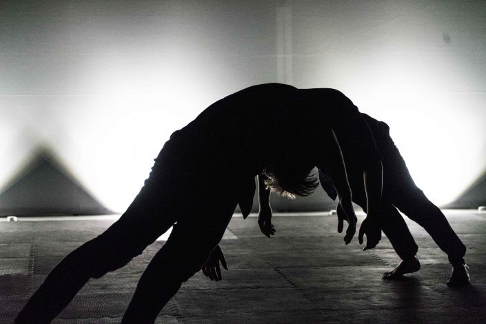 Two performers arch backward in silhouette beneath bright stage light.